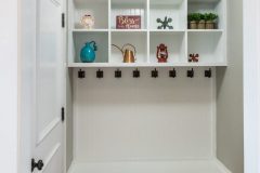 Hallway mud room in a new construction house home with white wooden shelves with decorative items, hooks for hanging jackets and a seat with storage baskets underneath