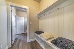 Mud room interior of a house with hooks shelves and built in bench with pillows. Wooden floor and open white doors can also be seen from the room.