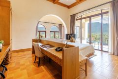 Luxury hotel suite interior features wooden workspace desk and bed overlooking balcony with mountain views. Mediterranean style decor with terracotta tiles and arched windows.